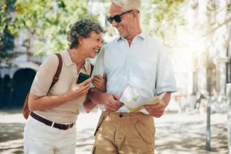 Elderly man and woman laughing. The man is holding a map, and the woman is wearing a backpack. They look like they are travelling and enjoying retirement.