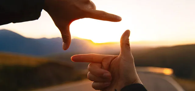 Hands framing a sunset over a winding road, symbolizing the importance of looking at the bigger picture when planning for retirement.
