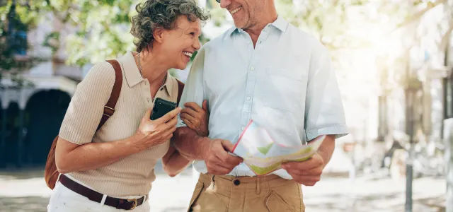 Elderly man and woman laughing. The man is holding a map, and the woman is wearing a backpack. They look like they are travelling and enjoying retirement.